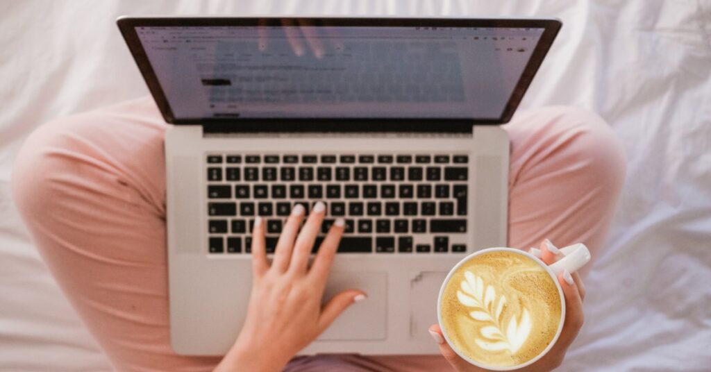 Woman working on laptop in pink pajamas drinking a latte